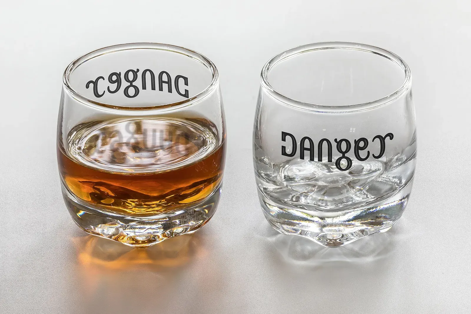 Ambigram Cognac / Danger , mirror symmetry (vertical axis), front and back, on a set of two shot glasses (full and empty). Humorous warning related t…